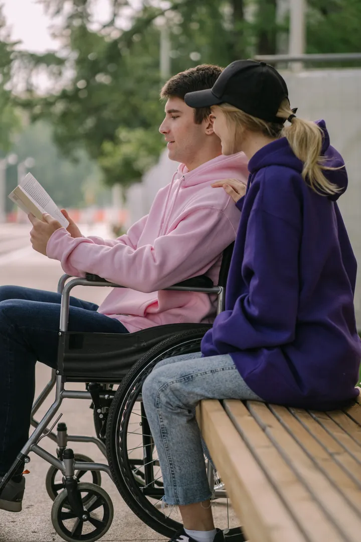 A young man in a wheelchair reading a book with a friend outdoors on a sunny day